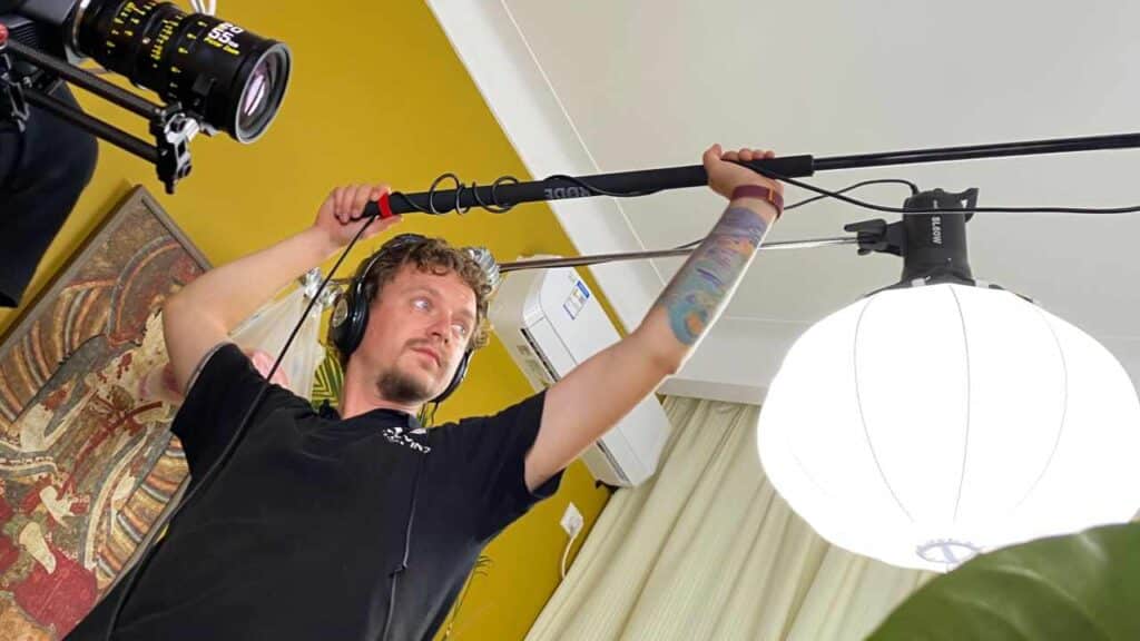 Professional sound recordist in Bangkok holding boom pole during indoor video interview setup with soft lighting and camera crew.